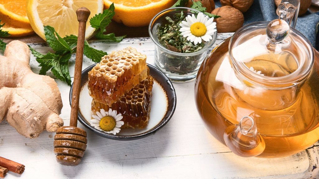 a table filled with an array of natural healthy foods to promote good health and wellness. the spread includes herbal tea in a glass tea pot, fresh ginger root, raw honey, citrus fruits and more